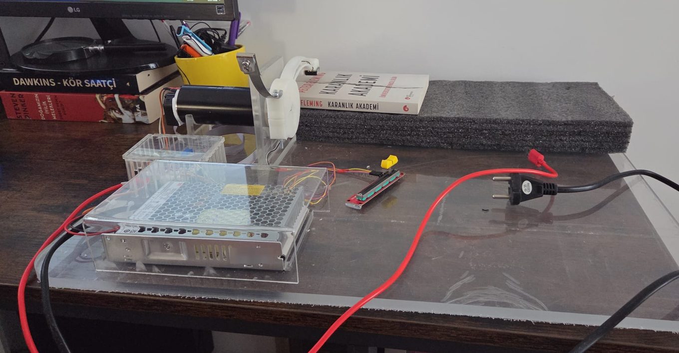 A force-matching rig on a desk. A metal power supply unit sits inside a clear acrylic enclosure on a transparent base plate, wired in red and yellow to a small controller board behind it. A black cylindrical motor is held in a white 3D-printed mount, with a curved metal lever arm rising from it. A slider potentiometer with a yellow thumb tab lies loose on the plate. Mains cables run off the desk, and books and a block of grey packing foam sit behind the rig.